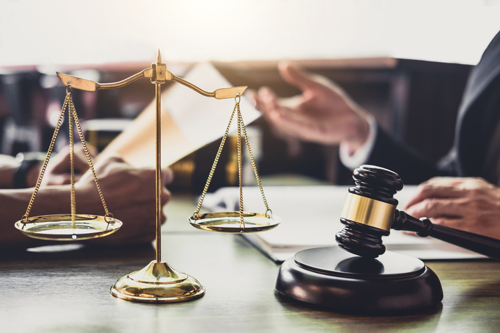Close-Up Of Weight Scale By Gavel With Lawyer And Client Discussing In Background