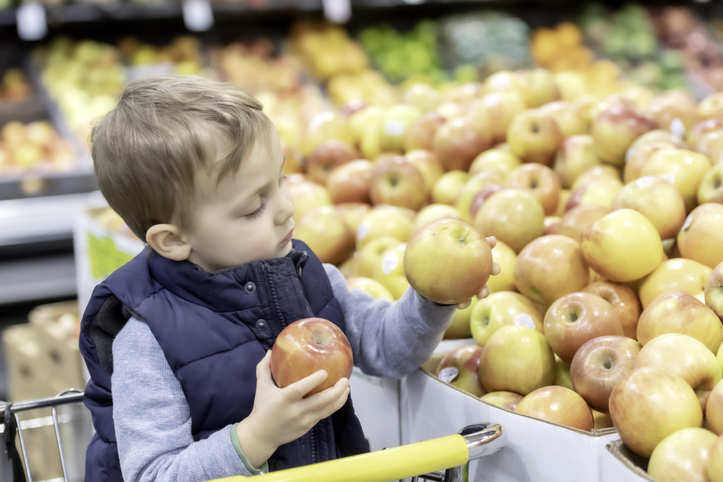 Three years old little child sitting in a shopping cart buying apples at a supermarket aisle