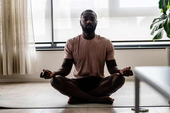 A young African-American man is meditating in a bright, minimalist living room, sitting cross-legged on the floor with eyes closed, practicing mindfulness and relaxation.