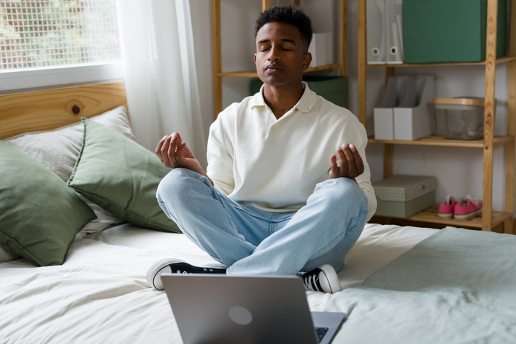 Young man practicing meditation on bed with laptop